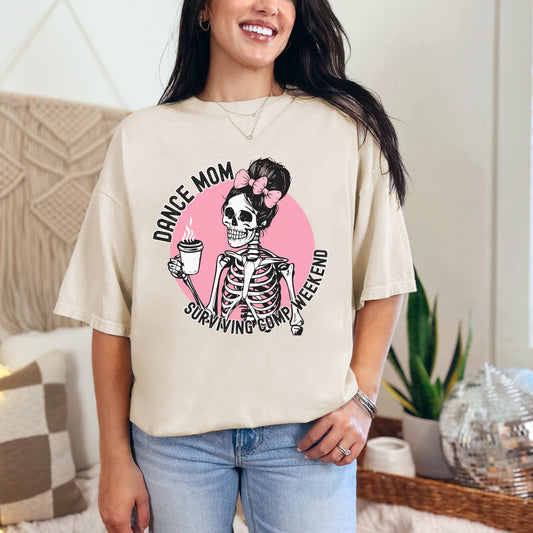 Trendy Comfort Colors Shirt with repeating words Dance Mom Surviving Comp Weekend circled around a messy bun pink bow coquette skeleton holding a cup of coffee with a pink background